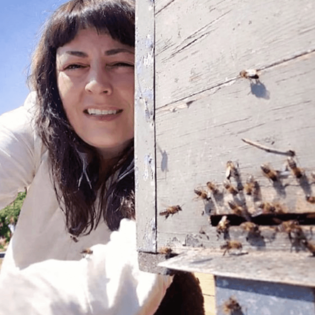 Una mujer sonriente se asoma detrás de una colmena de madera rodeada de abejas volando.