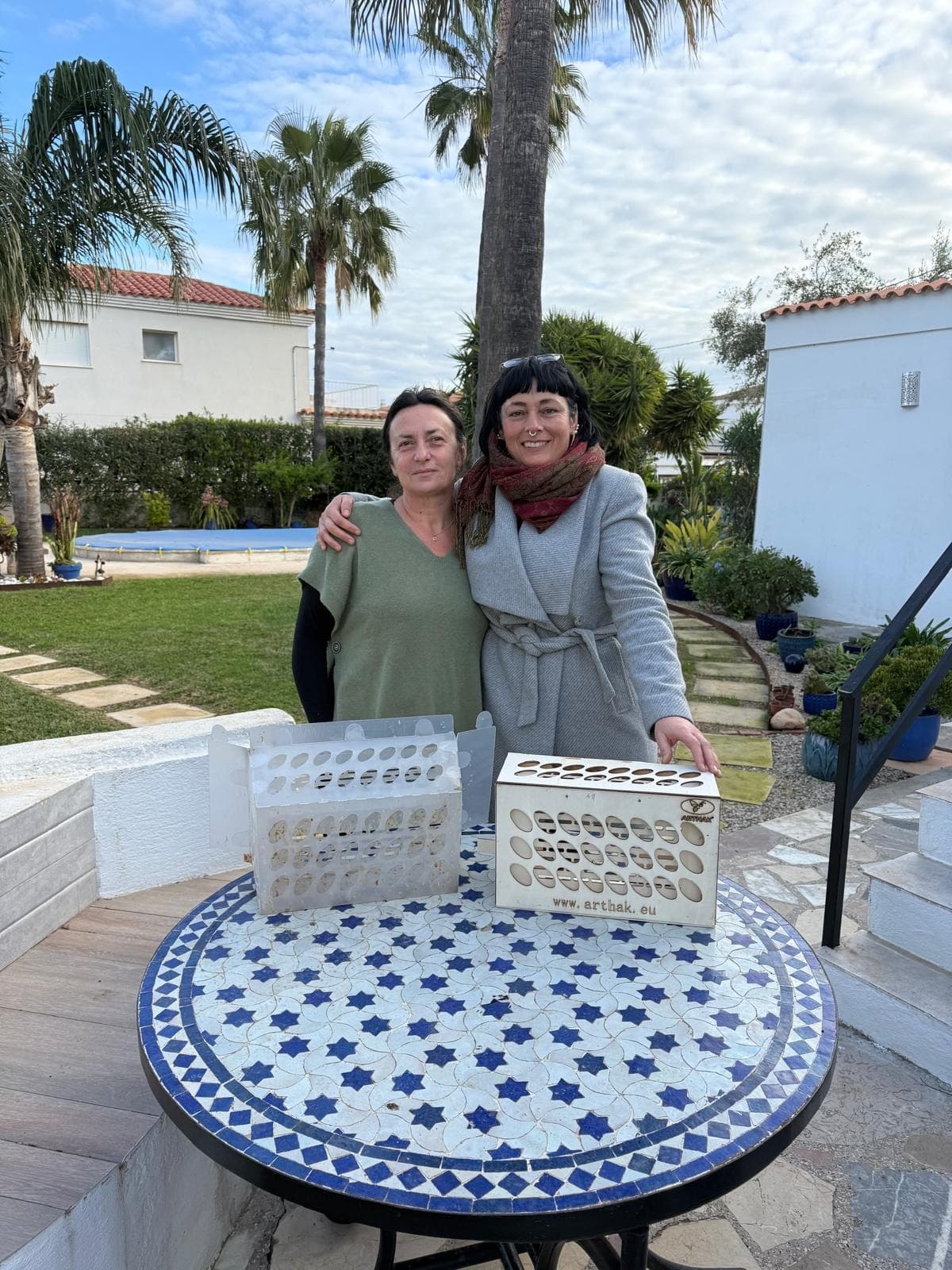 Two women pose behind a tiled table with two animal transport boxes in a garden.